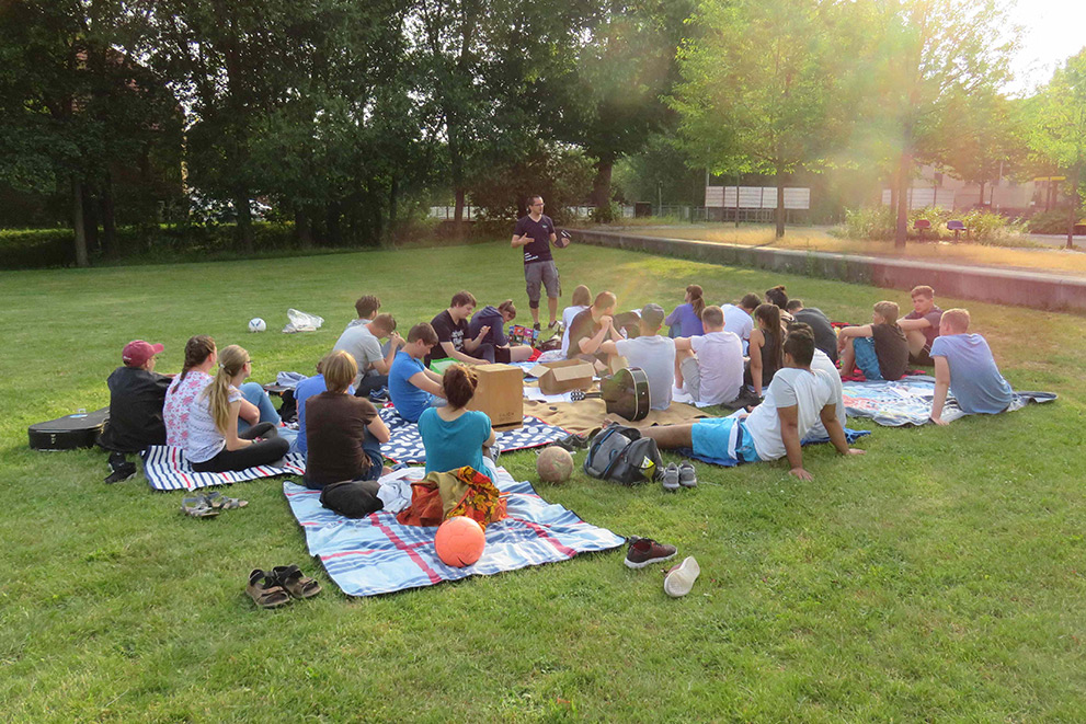Gruppe von Jugendlichen auf einer Wiese sitzend und dem Redner zuhören