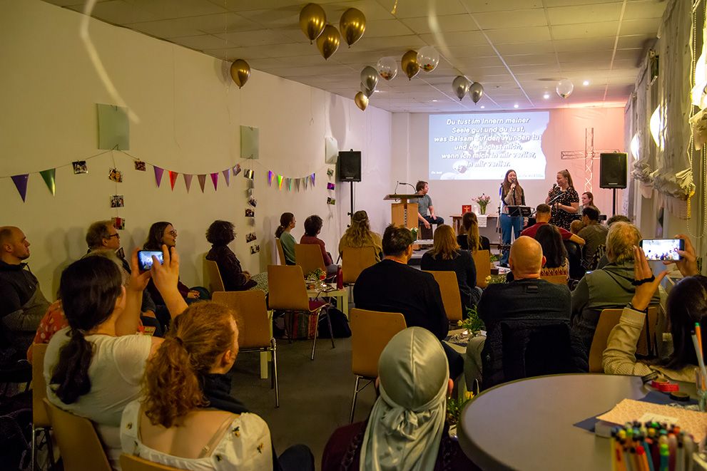 Größere Gruppe an Menschen in einem geschmückten Raum und 2 singende Mädchen im Vordergrund